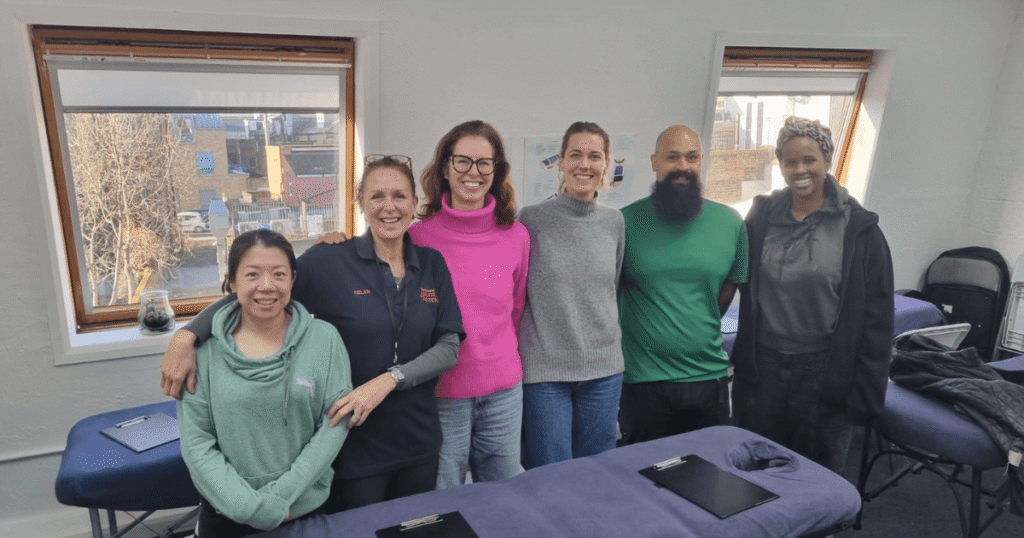 Helen Cameron-Lee standing with a group of massage students in a training classroom at The School of Natural Therapies, smiling beside massage tables during a practical session.